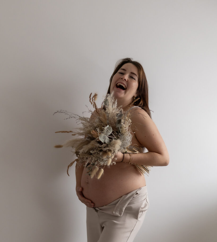 Schwangere Frau lacht mit einem Trockenblumenstrauß in der Hand & freiem Babybauch in die Kamera