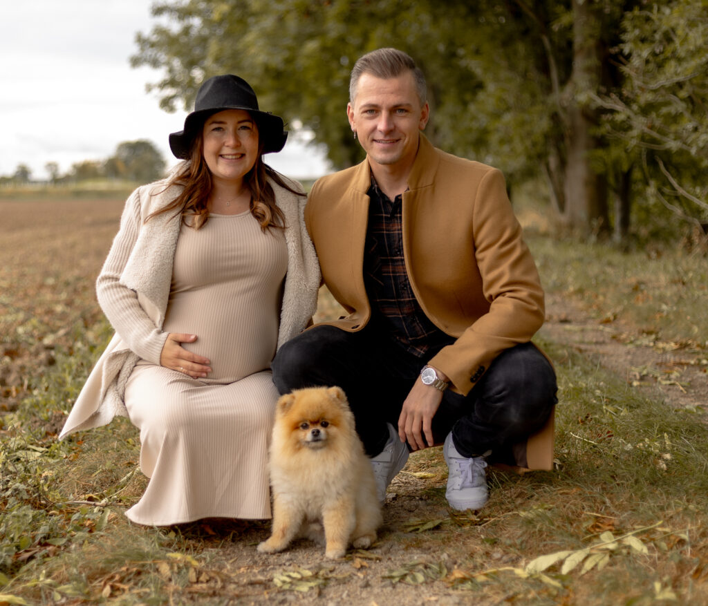 Schwangere Frau & Mann hocken hinter Hund auf dem Boden & schauen zur Kamera