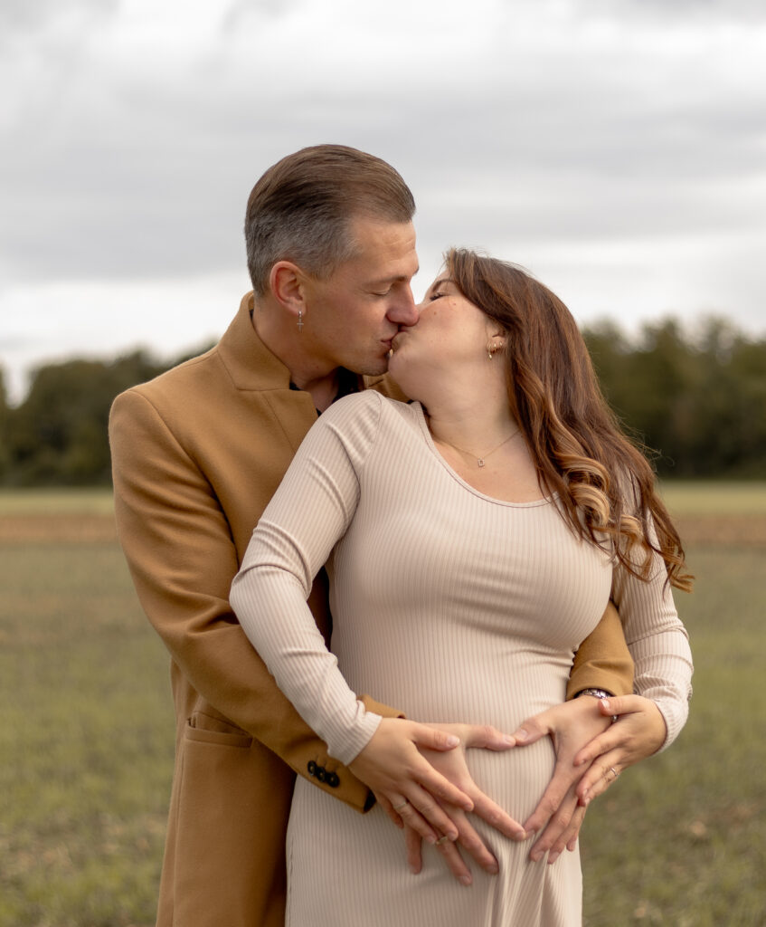 Schwangere Frau steht mit dem Rücken zu ihrem Mann, sie schaut nach hinten & beide küssen sich.