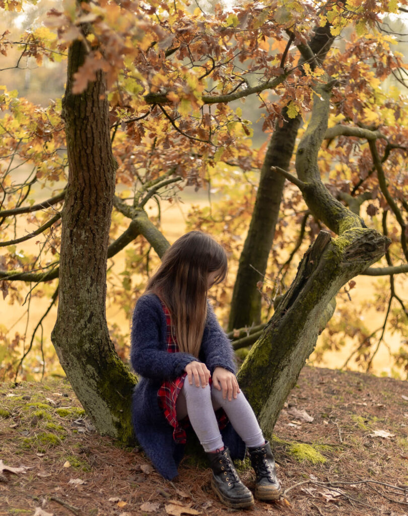 Tochter sitzt zwischen ein paar Baumstämmen & schaut nach hinten von der Kamera weg