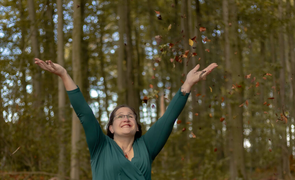 Nahaufnahme einer Frau, die auf dem Waldboden sitzt & Blätter in die Luft wirft