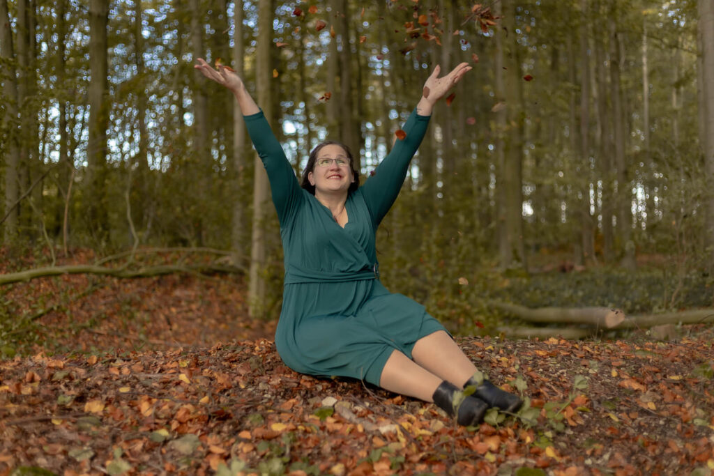 Frau sitzt auf dem Waldboden & wirft Blätter in die Luft
